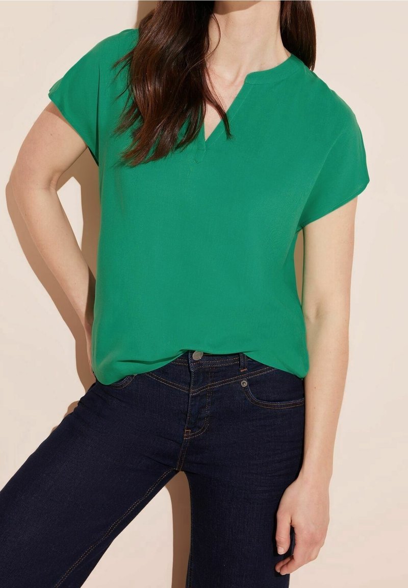 Donna che indossa una blusa verde con maniche corte e scollatura a V, insieme a jeans blu scuro, in piedi con una mano sui fianchi su uno sfondo beige.