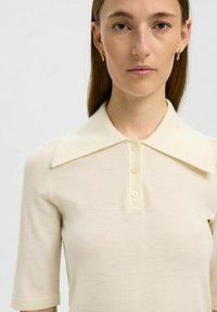 Camisa polo color crema con un cuello ancho, mangas cortas y cinco botones. La tela parece suave y blanda, con una textura ligera.