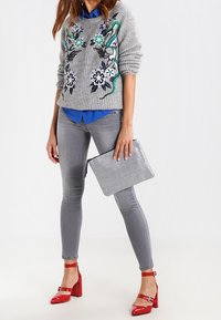 Pull gris en maille avec broderie florale, chemise bleue en dessous, jean skinny gris, et chaussures rouges à talon bloc verni, tenant une pochette argentée.