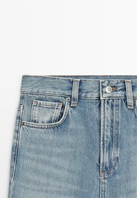 Jean en denim bleu clair à la taille avec passants pour ceinture, fermeture par bouton et un petit détail de poche avant sur fond blanc.