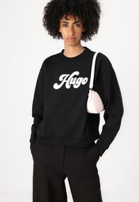 Felpa nera con logo bianco "Hugo". Vestibilità ampia, scollatura rotonda, polsini a coste. Abbinata a una piccola borsa rosa. Pantaloni neri a gamba larga.