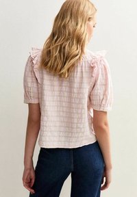 Rosa karierte Bluse mit Rüschensäumen, strukturiertem Stoff und lockerer Passform. Getragen mit dunkelblauen Jeans.
