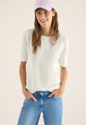 Kobieta uśmiechająca się, ubrana w jasnofioletową czapkę, biały teksturowany top z krótkim rękawem oraz niebieskie dżinsy, z rękami w kieszeniach, stojąca na jednolitym tle.