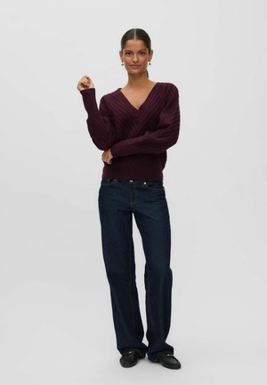 Kvinde iført en mørk lilla kabelsticket V-hals sweater, mørkeblå vidde-bukser og sorte loafers, stående med den ene arm hævet.