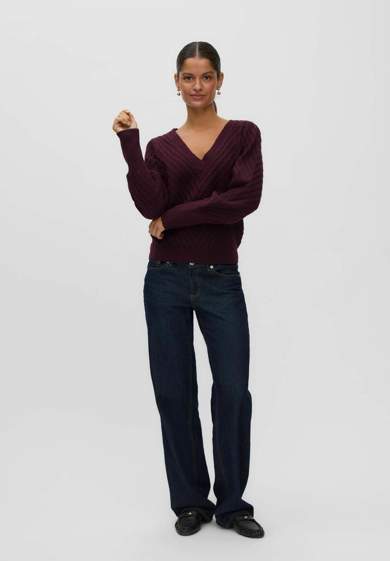Femme portant un pull en maille torsadée violet foncé à col en V, un jean large bleu foncé et des mocassins noirs, debout avec un bras levé.