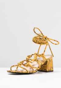 Żółte sandały na blokowym obcasie w wężowy wzór, z krzyżującymi się paskami i wiązaniami w okolicy kostki. Charakteryzują się fakturowaną powierzchnią i otwartym noskiem.