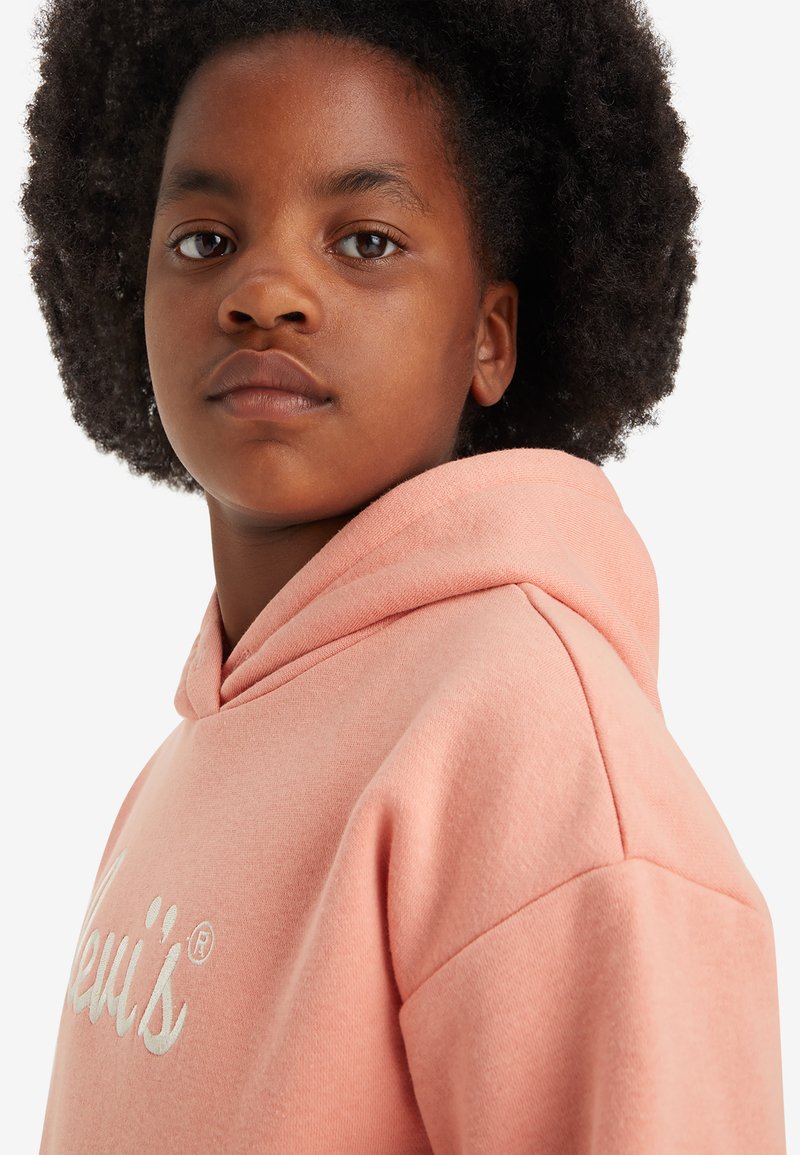 Roze hoodie met een zachte textuur, voorzien van een wit geborduurd logo op de borst, en een capuchon met trekkoord. Close-up van de halslijn en schouder.