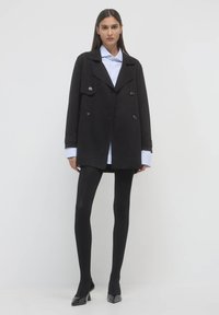 Cappotto nero doppiopetto con grandi bottoni, vestibilità rilassata, abbinato a una camicia a righe blu chiaro e collant neri. Tacchi neri lucidi.