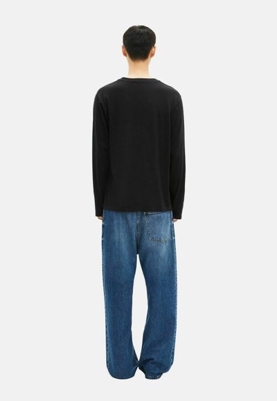 Persona de pie de espaldas, vistiendo una camiseta negra de manga larga y jeans de mezclilla azul holgados, contra un fondo blanco liso.