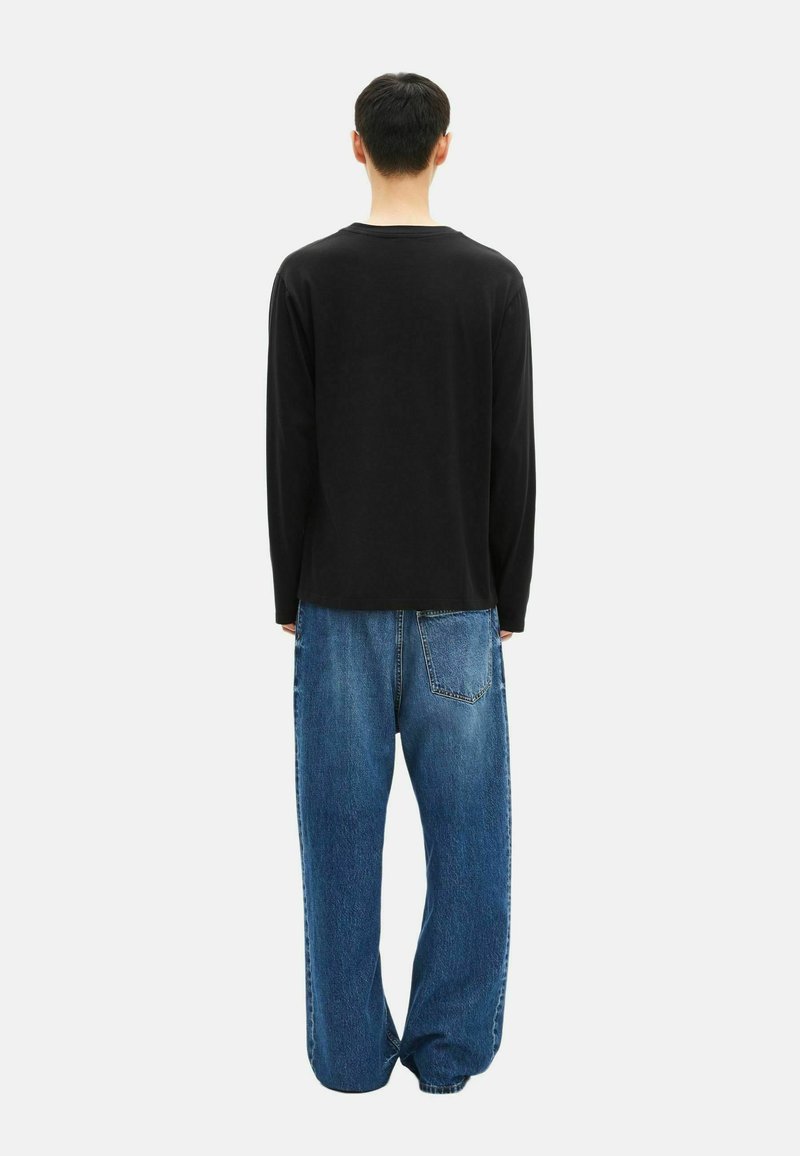 Persona in piedi di spalle, che indossa una maglietta nera a maniche lunghe e jeans in denim blu larghi, su uno sfondo bianco semplice.