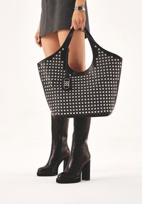 Sac tote noir avec des clous métalliques, grande ouverture en haut et un accent en matériel central. Texture lisse avec des larges sangles de shoulder.