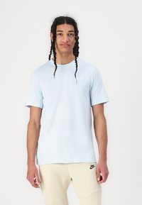 T-shirt bleu clair à manches courtes avec un col rond et un petit logo blanc sur la poitrine gauche. Porté avec un pantalon de sport beige.