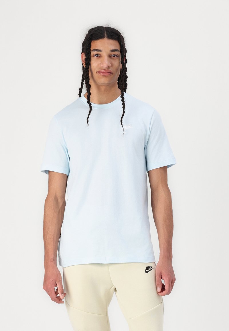 T-shirt bleu clair à manches courtes avec un col rond et un petit logo blanc sur la poitrine gauche. Porté avec un pantalon de sport beige.