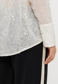 White sequined blouse with a sheer texture, featuring detailed patterns and an opaque white cuff. Worn over black trousers with side accents.