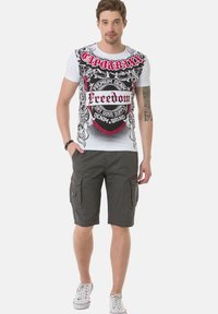 Witte t-shirt met gedurfde zwarte en rode grafische tekst en ontwerpen, gecombineerd met grijze cargoshorts met meerdere zakken. Witte sneakers.