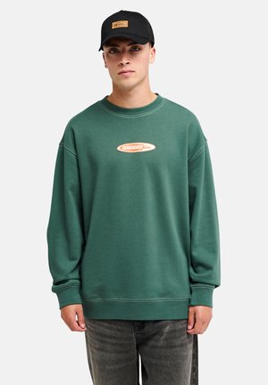 Mann trägt einen grünen Quiksilver-Sweatshirt, schwarze Kappe und ausgewaschene schwarze Jeans, steht vor einem einfarbigen hellen Hintergrund.