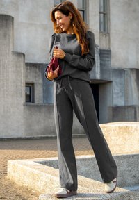 Grijze coördinateset bestaande uit een langemouwen top en een broek met wijde pijpen, met een trekkoord in de taille. Draagt met lichtgekleurde sneakers en een handtas.