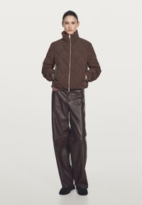 Braune steppte Jacke mit hohem Kragen und Reißverschluss auf der Vorderseite, kombiniert mit lockeren, glänzenden braunen Lederhosen, vor einem schlichten weißen Hintergrund.
