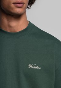 Mörkgrön bomullströja med rund hals, med en liten broderad "Westbury"-logotyp i vitt på vänster bröstområde. Slät textur.