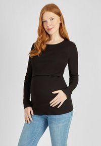 Fekete, hosszú ujjú kismama blúz texturált bordás mintával és ívelt aljjal, világoskék farmernadrággal párosítva. A modell az egyik kezét a pocakján tartja.