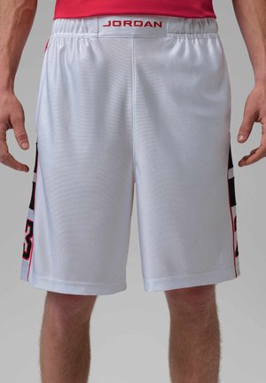 Weiße Basketball-Shorts mit rotem "JORDAN"-Label am elastischen Bund, schwarze und rote Seitenstreifen, getragen von einer Person mit sichtbaren Beinen und Händen.