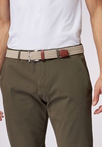 Geweven beige riem met een gestructureerd patroon, leren accenten in bruin en een zilveren metalen gesp, gedragen over olijfgroene broek.