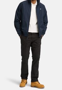 Mörkblå jacka med ståkrage, vit polo t-shirt, svarta byxor och gula skotertimbers-stil stövlar. Har en logotyp på jackan.