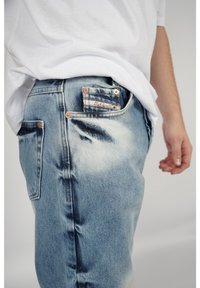 Jeans de mezclilla azul deslavada con un corte relajado, que cuentan con remaches de cobre, un bolsillo de parche y detalles de costura visibles en el costado.