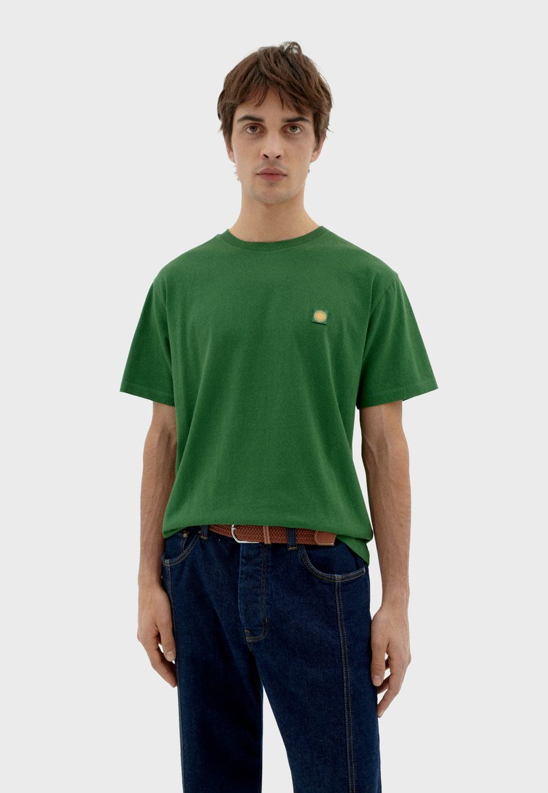 Camiseta de algodón verde con cuello redondo y mangas cortas, que presenta un pequeño logo de sol en el lado izquierdo del pecho. Llevada con jeans de color azul oscuro.
