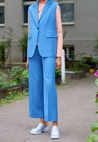 Blå ärmlös blazer och vida byxor med en rosa texturerad topp. Grå platåskor. Utomhusmiljö med grönska i bakgrunden.