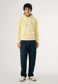 Felpa gialla in cotone con tasca a marsupio e lacci, abbinata a pantaloni blu navy morbidi e sneaker bianche con dettagli neri.