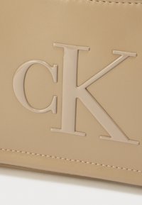 Beige läderväska med en stor, präglad "CK"-logotyp. Mjuk textur med subtila sömnadsdetaljer. Enkel, modern design.