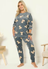 Harmaa fleece-pyjamakokonaisuus, jossa on pitkät hihat ja housut, ja jossa on toistuva nallekarhukuosi vaaleanbeigeinä. Pehmeä rakenne ja rento istuvuus.