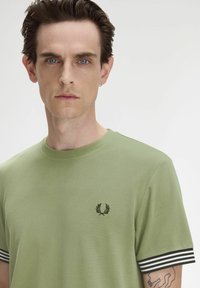 T-shirt in cotone verde con colletto a girocollo, caratterizzato da polsini a righe bianche e nere e un piccolo emblema ricamato sul petto.
