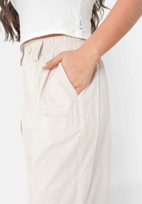 Beige relax-pantalon met een elastische tailleband, zijzakken en een subtiele geborduurde logo op het been. Glad fabricage textuur.