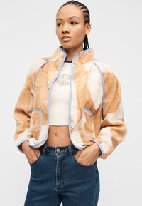 Γυναίκα με πλεξούδες φοράει πορτοκαλί και εκρού φλις μπουφάν με σχέδιο πάνω από λευκό crop top με γραφικό ήλιου, συνδυασμένο με μπλε τζιν.