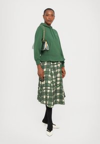 Felpa verde con taschino frontale, abbinata a una gonna scozzese verde e crema. Il modello indossa collant neri e tacchi bianchi con dettagli a contrasto.
