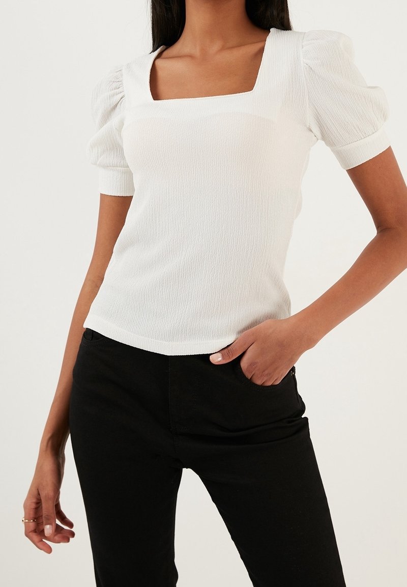 Biała, teksturowana bluzka z kwadratowym dekoltem i bufkami, zestawiona z dopasowanymi czarnymi spodniami. Kluczowe cechy to prążkowany materiał i krótkie rękawy.