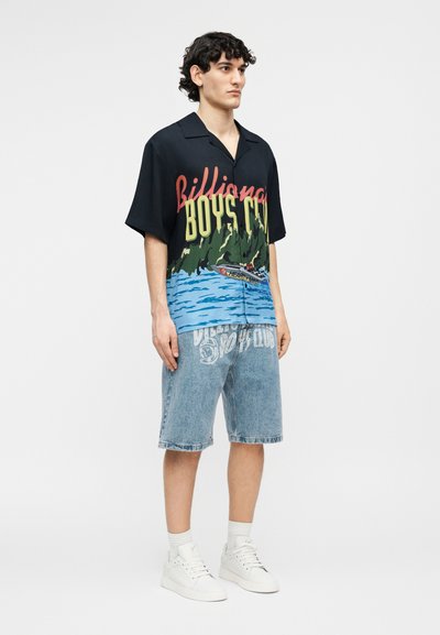 Νέος άνδρας που φοράει μαύρο κοντομάνικο με γραφικά και το κείμενο "Billionaire Boys Club", τζιν σορτς, λευκές κάλτσες και λευκά αθλητικά παπούτσια.