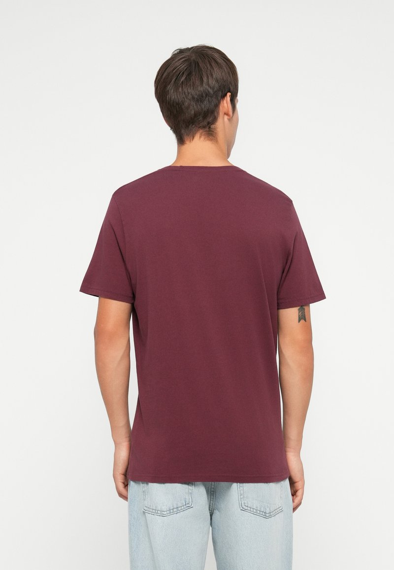 Uomo con capelli corti castani che indossa una semplice maglietta bordeaux e jeans azzurri chiari, di spalle e su uno sfondo bianco.