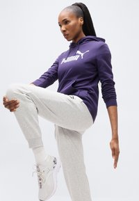 Μωβ φούτερ με κουκούλα και λευκό λογότυπο, γκρι φόρμα και λευκά παπούτσια. Το σύνολο έχει casual σχεδιασμό κατάλληλο για αθλητική χρήση.