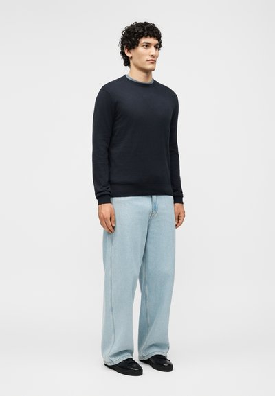 Άνδρας που στέκεται φορώντας μαύρο πουλόβερ με μακριά μανίκια, ανοιχτό μπλε παντελόνι φαρδιάς γραμμής και μαύρα παπούτσια μπροστά σε απλό λευκό φόντο.