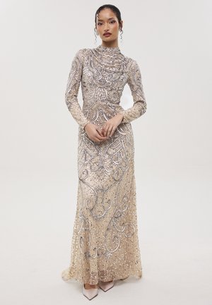 Mujer con un vestido beige de manga larga y largo hasta el suelo, adornado con intrincados abalorios plateados y lentejuelas, de pie frente a un fondo claro y simple.