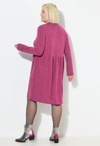 Fuchsia jurk met lange mouwen en uitlopende onderkant, tot over de knie, gemaakt van een gestructureerde stof. Gecombineerd met metallic enkellaarsjes met roze accenten.