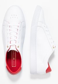 Un par de zapatillas bajas de cuero blanco con cuello acolchado rojo y ojales dorados, plantillas de marca y detalles de perforación en la puntera.