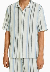 Camisa de botones de manga corta hecha de una tela ligera con rayas verticales en azul claro, negro y crema. Textura suave, corte relajado.