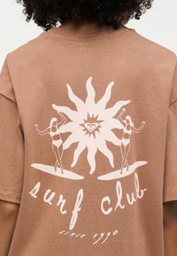 Καφέ μπλουζάκι με λευκό γραφικό δύο γυναικών σε σανίδες σερφ που πλαισιώνουν έναν στυλιζαρισμένο ήλιο, κείμενο «surf club since 1990» από κάτω.