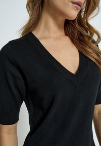 Top en tricot noir avec un large décolleté en V, des manches courtes et une silhouette ajustée. Le tissu a une texture lisse et un léger stretch.