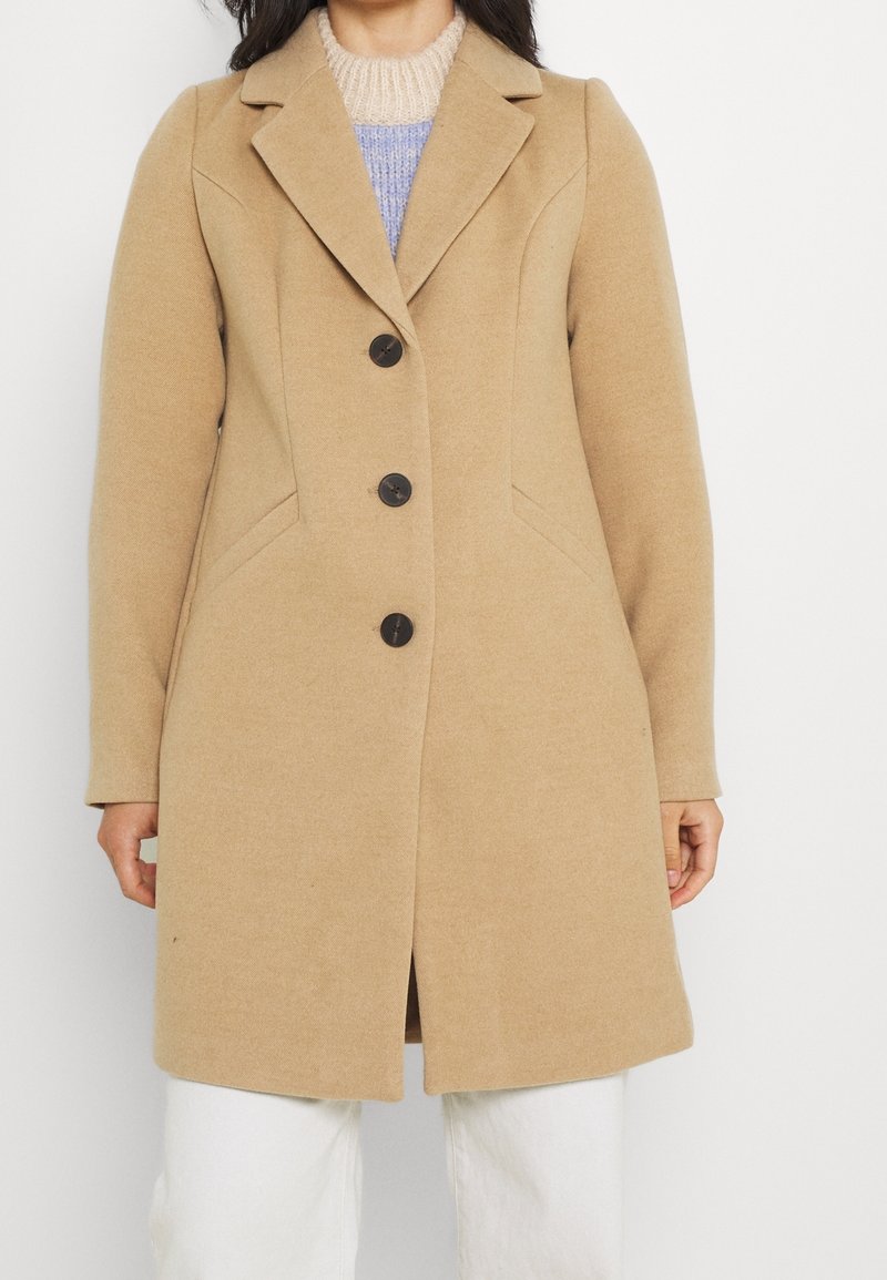Abrigo de lana de camello con un diseño ajustado, solapas de muesca, tres botones oscuros y bolsillos laterales. Textura suave y corte a la altura de la rodilla.