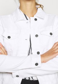 Veste en denim blanc avec des boutons noirs, deux poches poitrine, un col et des manches longues. Tissu texturé avec un design épuré.